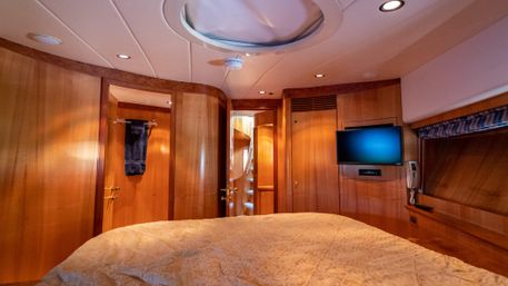 Cozy wood-paneled yacht cabin bedroom with quilted bed, overhead hatch, wall-mounted TV and open doorway to stairs