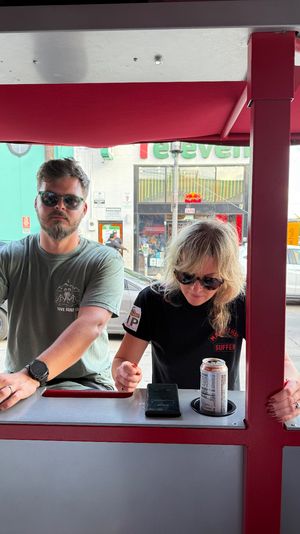 Two people wearing sunglasses at a red outdoor counter under an awning, opening canned drinks and checking a phone with downtown storefronts and parked cars visible on the street behind them.