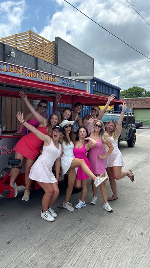 Cheerful group of women in pink and white dresses posing and waving on a red party pedal bike parked on an urban street by industrial buildings under a partly-cloudy sky