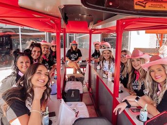 Smiling group of women in pink cowboy hats and matching shirts riding a red pedal-pub party bike down a sunny downtown street.