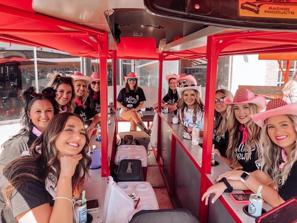 Smiling group of women in pink cowboy hats and matching shirts riding a red pedal-pub party bike down a sunny downtown street.