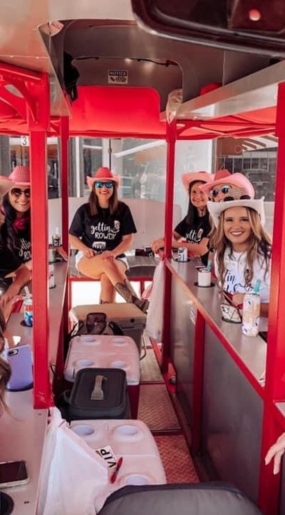 Smiling group of women in pink cowboy hats and matching shirts riding a red pedal-pub party bike down a sunny downtown street.
