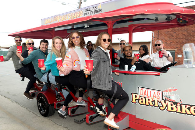 Smiling group of people on a red pedal-powered party bike on a downtown street, holding red cups and enjoying a daytime ride.