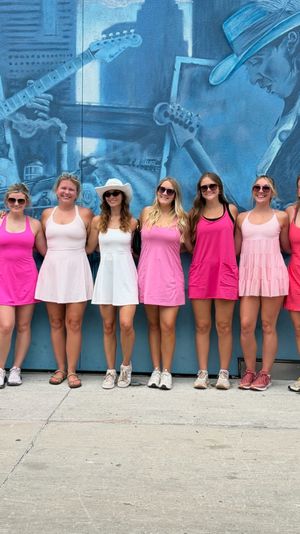 Group of women in pink and white summer dresses posing on a downtown sidewalk in front of a large blue music-themed mural depicting guitarists.