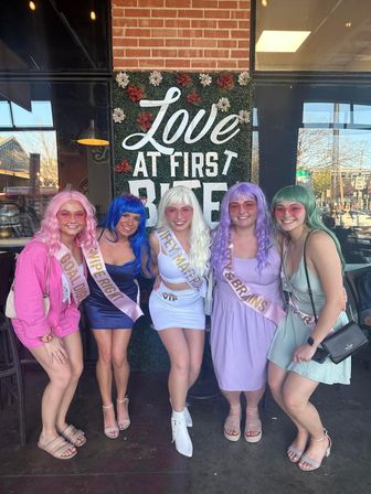 Five women in bright wigs and sashes pose on a restaurant patio beneath a floral "Love at First Bite" sign — colorful bachelorette brunch scene.