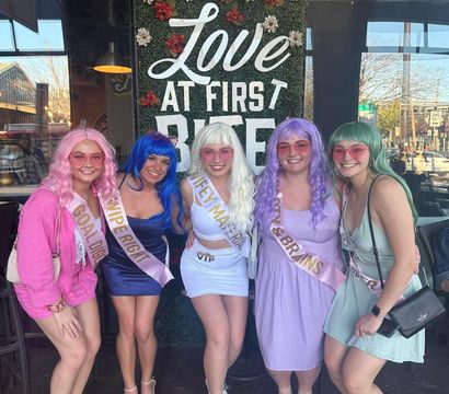 Five women in bright wigs and sashes pose on a restaurant patio beneath a floral "Love at First Bite" sign — colorful bachelorette brunch scene.
