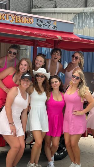Group of women in pink and white dresses smiling and posing on and around a red pedal-powered party bike parked on a Dallas street, colorful mural and Jeep in the background.