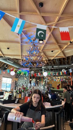 Bartender pouring a cocktail behind a busy bar in a festive sports-bar interior, wood coffered ceiling with antler chandelier, strings of international flags, hanging globe lights and patrons in the background.