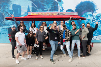 Cheerful group posing around a red pedal-powered party bike with a canopy in front of a vibrant blue music-themed mural on a Dallas sidewalk.