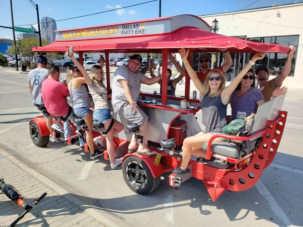 Group of friends riding a red open-air pedal party bike on a sunny Dallas street, smiling and cheering with downtown skyline visible