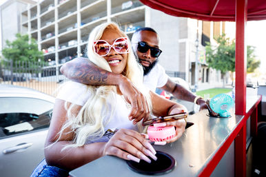 Smiling couple in sunglasses leaning at an outdoor downtown drink cart on a city street; man with tattooed arm hugs woman with long blonde hair as she holds a phone and a pink handheld fan, parking garage in the background.