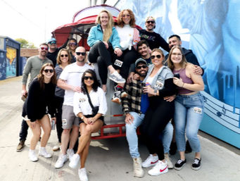 Smiling group of adults posing on and around a red trolley-style vehicle beside a blue mural on a sunny urban sidewalk.
