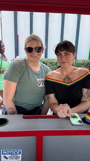 Two friends smiling at a red booth during an outdoor Pride street festival on a city sidewalk — one in sunglasses with a VIP sticker, the other in a black off-shoulder top with rainbow trim and tattoos, crowd and staff behind them.