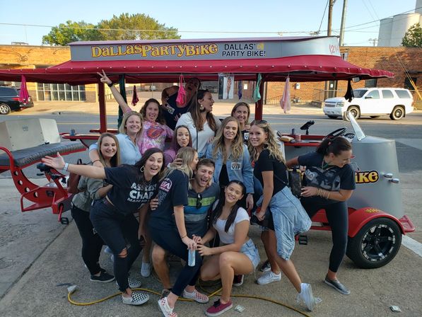Large group of friends laughing and posing on a red party bike pedal pub parked on a sunny downtown street with hanging decorations and drinks.