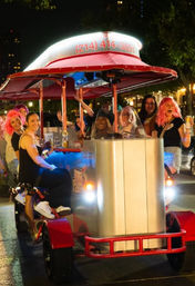 Group of friends enjoying a brightly lit pedal pub on a downtown street at night, laughing, holding drinks and wearing colorful wigs.