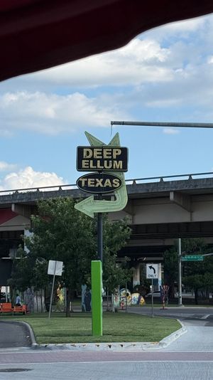 Neon arrow sign reading "Deep Ellum Texas" pointing left beneath an overpass in Deep Ellum, Dallas, Texas, with blue sky and street scene