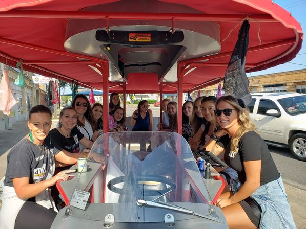 Group of smiling women riding a red-canopied pedal bar (party bike) with drinks, cruising a sunny downtown street by storefronts.