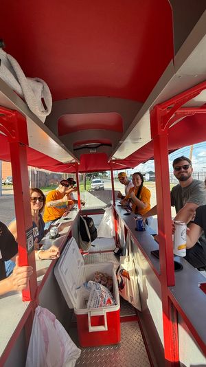 Smiling group riding a red-covered pedal pub party bike, holding drinks around a central cooler during a sunny urban street tour