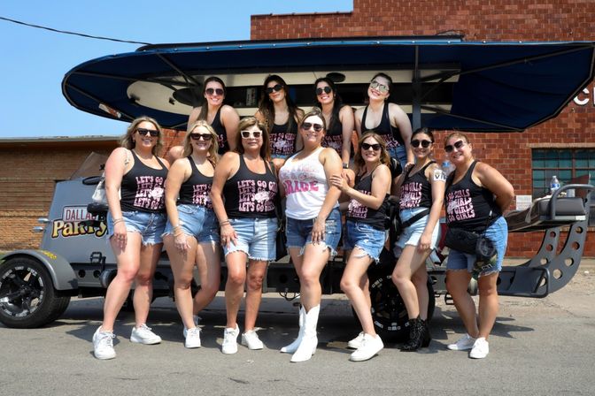 Bachelorette party group of women in matching "Let’s Go Girls" black tanks and denim shorts with the bride in a white tank and white cowboy boots, all wearing sunglasses and posing in front of an open-air party vehicle on a sunny urban street.