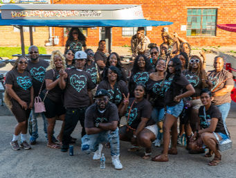 Large group of friends and family posing on a sunny urban street, many wearing matching black t‑shirts with teal heart design, gathered in front of a brick building and food tent, smiling and striking playful poses