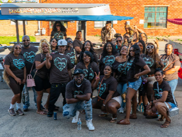 Large group of friends and family posing on a sunny urban street, many wearing matching black t‑shirts with teal heart design, gathered in front of a brick building and food tent, smiling and striking playful poses
