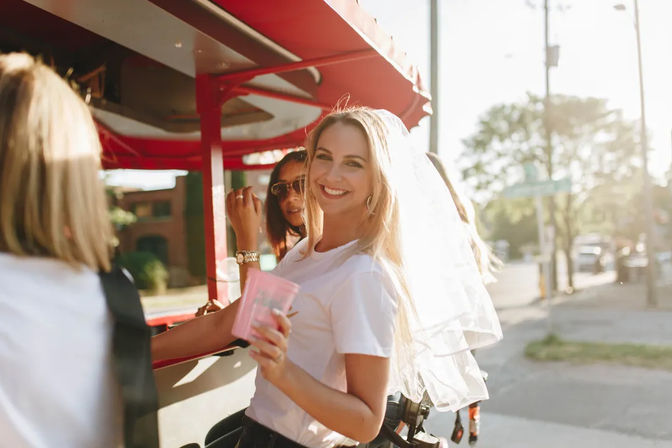 Bride-to-be wearing a veil laughs with friends on a sunny urban pedal party trolley, holding a pink cup during a daytime bachelorette outing.