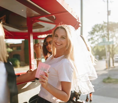 Bride-to-be wearing a veil laughs with friends on a sunny urban pedal party trolley, holding a pink cup during a daytime bachelorette outing.