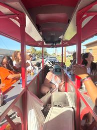 Group of people on a red-canopy party pedal bike cruising a sunny small-town street, passengers sipping drinks and grabbing cans from coolers down the center aisle.
