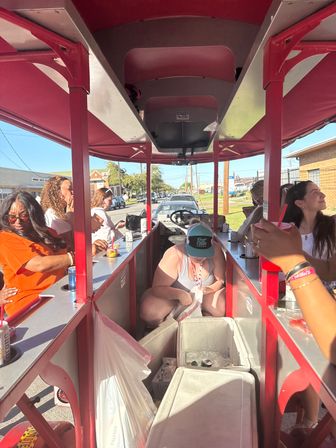 Group of people on a red-canopy party pedal bike cruising a sunny small-town street, passengers sipping drinks and grabbing cans from coolers down the center aisle.