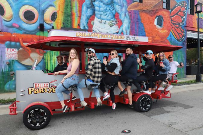 Group of people riding a red pedal-powered party bike on a Dallas street, smiling and holding drinks in front of a vibrant, colorful mural.