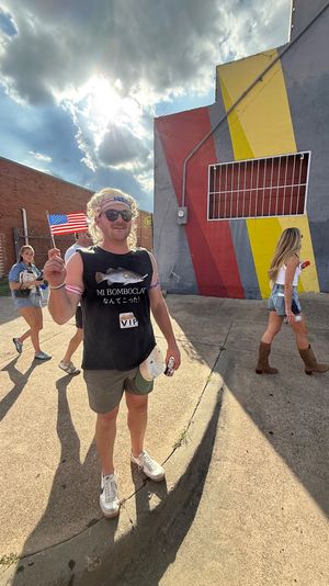 Smiling person in a sleeveless shirt and headband waving a small American flag on a sunny urban sidewalk in front of a colorful geometric mural, with other festival-goers walking by.