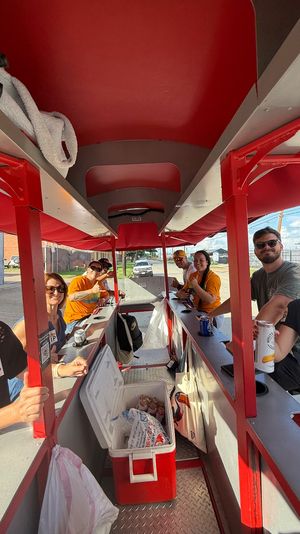 Smiling adults riding a red pedal-pub party bike with open-air bar seating, coolers and drinks while cruising a sunny urban street