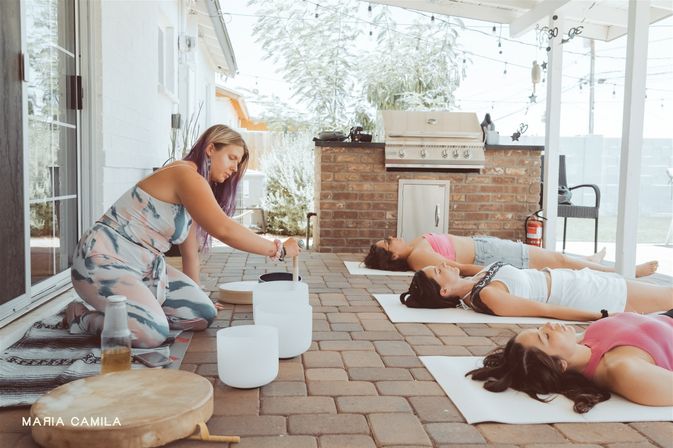 Woman playing crystal singing bowls for a relaxing outdoor sound bath on a covered backyard patio, while three people lie on yoga mats near a brick grill and string lights