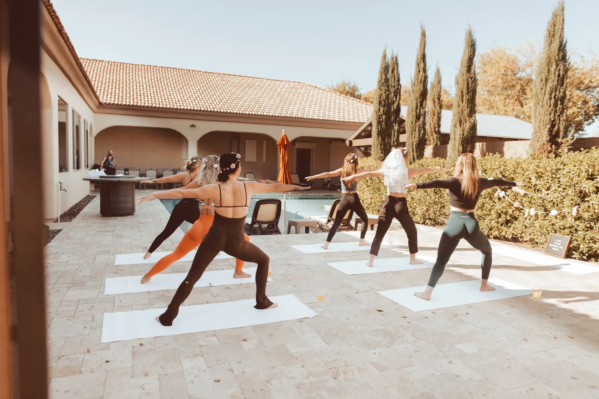 Group of women practicing outdoor poolside yoga on mats on a sunny tiled patio beside a Mediterranean-style house and tall cypress trees.