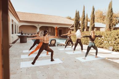 Group of women practicing outdoor poolside yoga on mats on a sunny tiled patio beside a Mediterranean-style house and tall cypress trees.