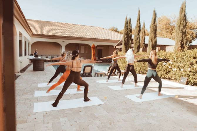 Group of women practicing outdoor poolside yoga on mats on a sunny tiled patio beside a Mediterranean-style house and tall cypress trees.