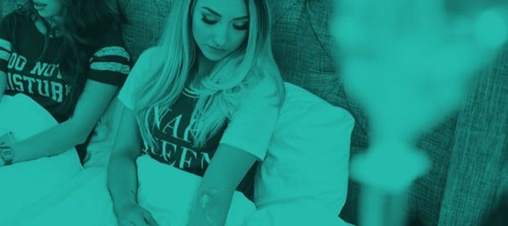 Two women lounging on a bed with pillows in a cozy bedroom, one applying lotion to her arm while the other sits beside her; teal-tinted, blurred foreground.