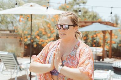 Woman in sunglasses and patterned cover-up making a namaste gesture poolside in a sunny backyard with umbrellas, lounge chairs and orange flowering shrubs