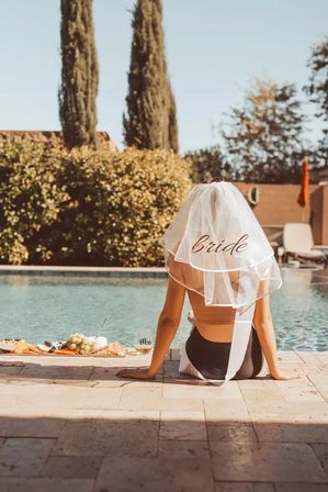 Bride veil poolside — woman sitting at a sunny backyard pool with her legs in the water, charcuterie board and drink beside her, tall cypress trees in the background.