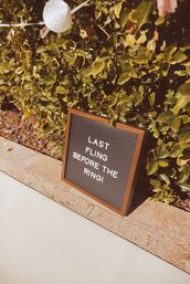 Sunlit letter-board sign reading 'Last fling before the ring!' leaning against a leafy hedge on a stone patio, white paper lantern and party decor suggesting an outdoor bachelorette celebration