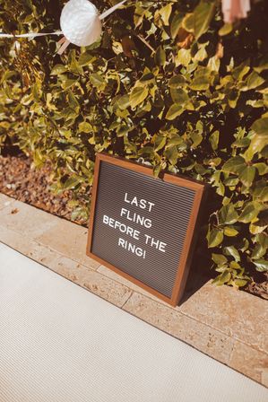 Sunlit letter-board sign reading 'Last fling before the ring!' leaning against a leafy hedge on a stone patio, white paper lantern and party decor suggesting an outdoor bachelorette celebration