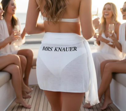Bachelorette boat party: woman in a white bikini and sheer sarong with black lettering stands on a yacht deck holding champagne, surrounded by laughing friends in white.