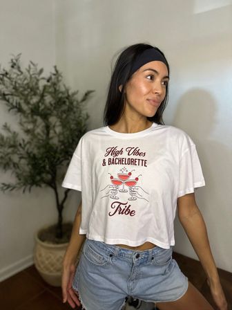 Woman in a black headband wearing a white cropped T-shirt printed 'High Vibes & Bachelorette Tribe' and denim shorts, posing indoors beside a potted plant — playful lifestyle product shot.