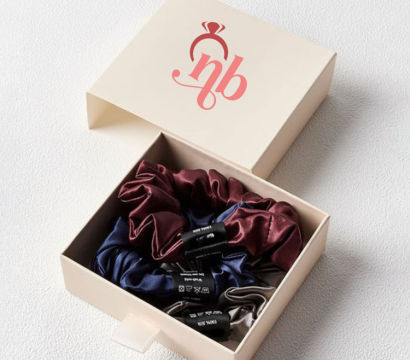Open cream gift box with three satin hair scrunchies — burgundy, navy, and gray — nestled on a textured white surface.