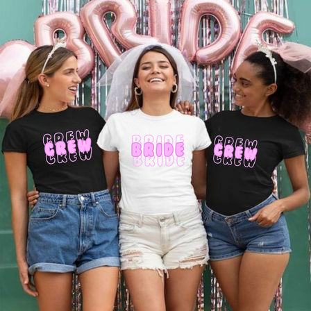 Bachelorette party: three women smiling under rose-gold 'BRIDE' balloons — bride in white 'BRIDE' tee and veil, two friends in black 'CREW' tees and denim shorts.