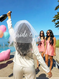 Poolside bachelorette party — woman wearing a "bride to be" veil raises a champagne bottle while two friends in pink satin dresses smile by the ocean with pastel balloons and a clear blue sky