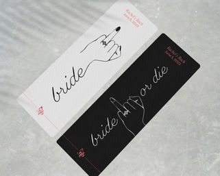 Two diagonal black and white bachelorette party cards on a marble surface, minimalist script “bride” design with a playful line-art hand flipping the middle finger wearing an engagement ring — sassy bridal shower keepsake