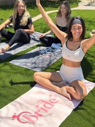 Three women in activewear enjoying an outdoor yoga session on a sunny grass lawn, smiling and stretching on yoga mats including a pink mat labeled "Bride" for a relaxed group fitness/wellness vibe.