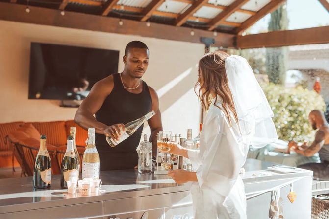 Bride-to-be in a white robe and veil at a sunny outdoor patio bar as a bartender pours champagne into her glass, several champagne bottles lined on the counter — bachelorette celebration vibe.