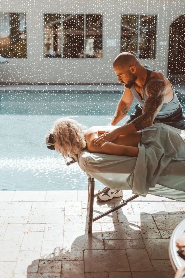 Tattooed male masseur giving an outdoor poolside back massage to a woman on a portable table beside a sunlit resort-style pool, spa relaxation scene.